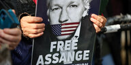 Eine Person hält ein Plakat mit dem Gesicht von Assange darauf. Übe rseinen. Lippen eine US-Fahne montiert. Darunter steht "Freee Assange"-