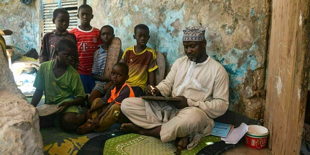 Der Lehrer Abas M‘Ballo sitzt in heller traditioneller Kleidung mit einer Tafel auf dem Boden, neben ihm sitzen sechs Jungen, die ihm zuhören