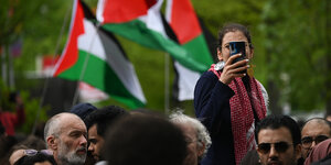 Eine Frau mit Smartphone sitzt vor der Veranstaltungshalle des Palästina-Kongress 2024 vor Palästinafahnen auf Schultern.